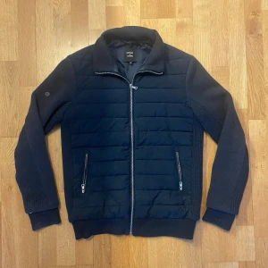 Mörkblå jacka från Superdry Studios - Snygg mörkblå cardigan från Superdry med dun på framsida och dragkedja. Jackan har långa ärmar och ribbade muddar, perfekt för kyligare dagar. Den stilrena designen gör den lätt att matcha med olika outfits.