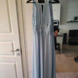 Ljusblå långklänning med spetsdetaljer - Elegant ljusblå långklänning med spetsdetaljer i midjan och en v-ringad design. Klänningen har en ärmlös stil och en mjuk, flödande passform som ger en romantisk känsla.