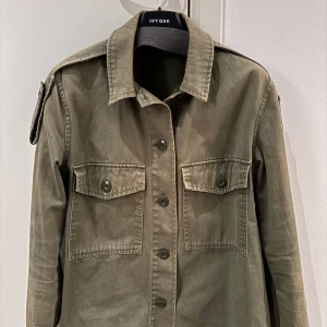 Grön jacka från Mango - Snygg grön overshirt/jacka från Mango. Jackan har två bröstfickor med lock och knappar, vilket ger en cool militärinspirerad look. Perfekt för lager-på-lager-stil under höst och vår.