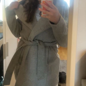 Grå kappa med knytband - Snygg grå kappa med knytband i midjan och knappar framtill. Perfekt för kyligare dagar med sin stilrena design och långa ärmar. Använd minst 3 gånger och är nästan helt ny. Inga fläkar eller nått