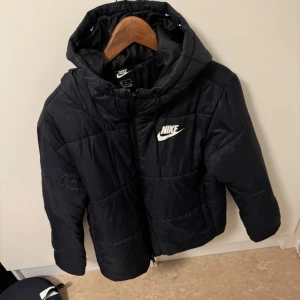 Svart dunjacka från Nike - Säljer en svart dunjacka från Nike med huva och dragkedja. Jackan har en klassisk design med Nike-logga på bröstet. Perfekt för kyliga dagar.