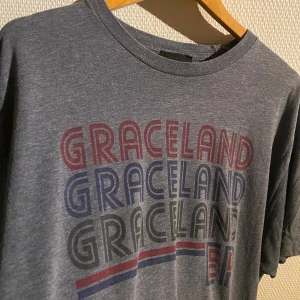 Grå t-shirt med Graceland-tryck - Snygg grå t-shirt med tryck av 'Graceland' i rött och blått. Perfekt för fans av Elvis Presley. T-shirten har en klassisk rund hals och korta ärmar.