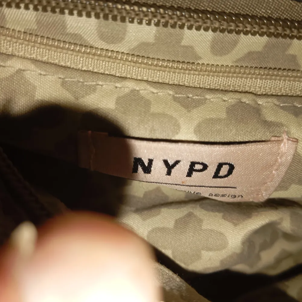 Säljer en snygg beige axelväska från NYPD. Väskan är i skinn och har en stilren design med en stor ringdetalj på framsidan. Insidan är mönstrad och har en praktisk dragkedja. Perfekt för både vardag och fest!. Väskor.