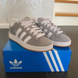 Gråa Adidas Campus sneakers - Säljer ett par snygga gråa Adidas Campus sneakers i nyskick. Skorna är gjorda av mocka och har vita detaljer som de klassiska tre ränderna och snörning. De kommer med extra gråa skosnören. Perfekta för både vardag och fest!
