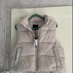 Beige dunväst med dragkedja - Säljer en snygg och bekväm beige dunväst. Den har en dragkedja framtill och två fickor med knappar. Perfekt för kyliga dagar när du vill hålla dig varm men ändå se stilren ut. Passar bra till både vardag och lite mer avslappnade tillfällen.