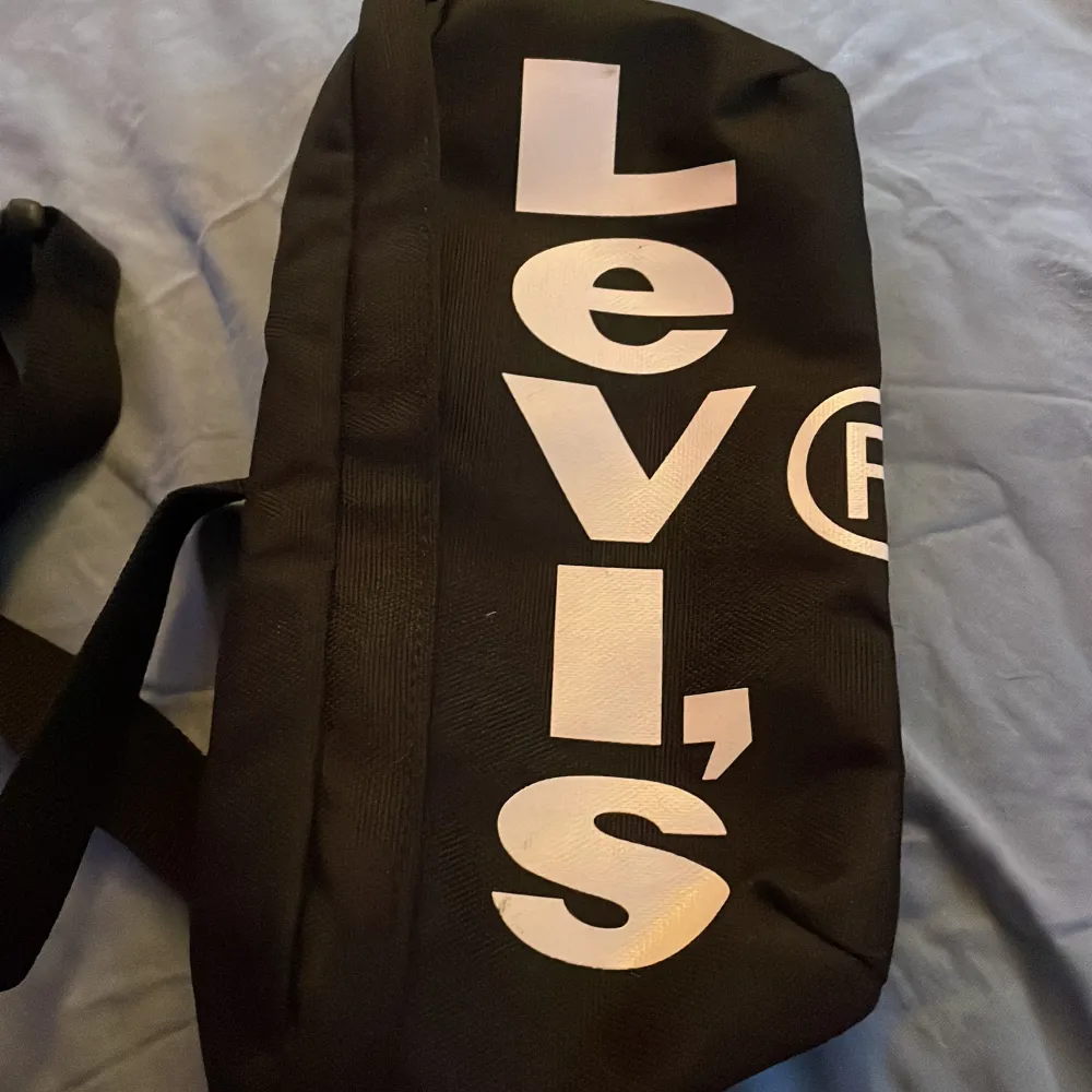 Säljer en snygg svart axelväska/magväska från Levi's. Väskan har en justerbar rem och ett stort vitt tryck med Levi's logga på framsidan. Perfekt för vardagsbruk och rymmer det mesta du behöver ha med dig. Den har en dragkedja för säker förvaring.. Väskor.