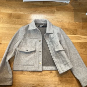 Grå ulljacka från Zara - Säljer en snygg grå kort kappa från Zara. Den har en klassisk krage och stängs med knappar framtill. Jackan har två stora fickor på bröstet som ger en cool look. Perfekt för höst och vår!