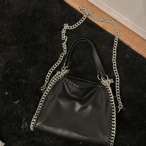 Svart handväska från Gina Tricot - Säljer en snygg svart handväska från Gina Tricot. Väskan har en kedja som axelrem och detaljer runt kanterna. Den är gjord av syntetmaterial och har en dragkedja på insidan för extra säkerhet. Perfekt för både vardag och fest!