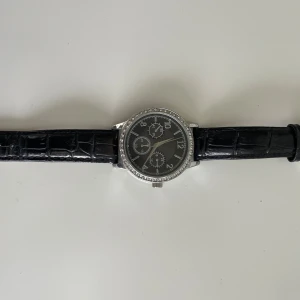 Svart klocka med silverdetaljer - Säljer en snygg klocka från Glitter med svart urtavla och silverdetaljer. Klockan har en svart rem i skinn med krokodilpräglat mönster. Runt urtavlan finns det glittriga stenar som ger en lyxig känsla. Klockan har tre små urtavlor inuti och är i nyskick. Perfekt accessoar för både vardag och fest! Batteriet behöver bytas. 