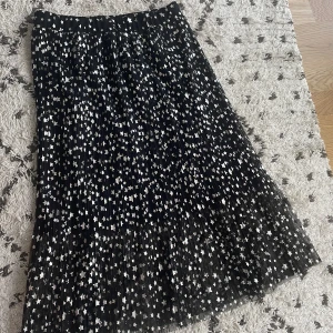 Svart kjol med stjärnmönster från H&M - Säljer en superfin svart kjol från H&M med ett coolt stjärnmönster i silver. Kjolen är plisserad och har en elastisk midja för extra komfort. Den är perfekt för både vardag och fest! Kjolen är i storlek M och är i mycket bra skick. Passar perfekt till höst och vinter outfits. 🌟