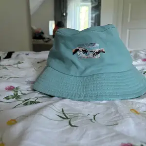 Säljer en ljusblå bucket hat med ett coolt broderi av två händer som nästan rör vid varandra. Hatten är perfekt för sommaren och har en avslappnad, normal passform. Materialet känns som bomull och är riktigt skönt att ha på sig. Perfekt för att skydda sig mot solen på stranden eller bara som en snygg accessoar till din outfit!