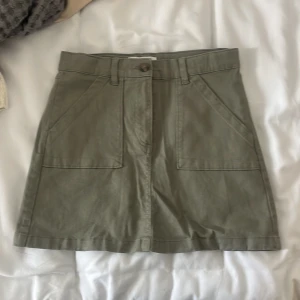 Olivgrön kortkjol - Säljer en superfin olivgrön miniskirt. Den har en enkel och stilren design med två stora fickor fram och en knapp- och dragkedjestängning. 