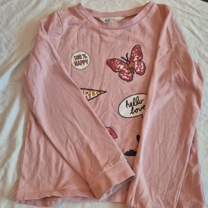 Rosa långärmad tröja från H&M - Säljer en söt rosa långärmad tröja från H&M i storlek 134/140. Tröjan har flera tryck på framsidan, inklusive en fjäril, en fågel, en stjärna, läppar och text som '100% Happy', 'Girls' och 'hello love'. Perfekt för höst och vår!