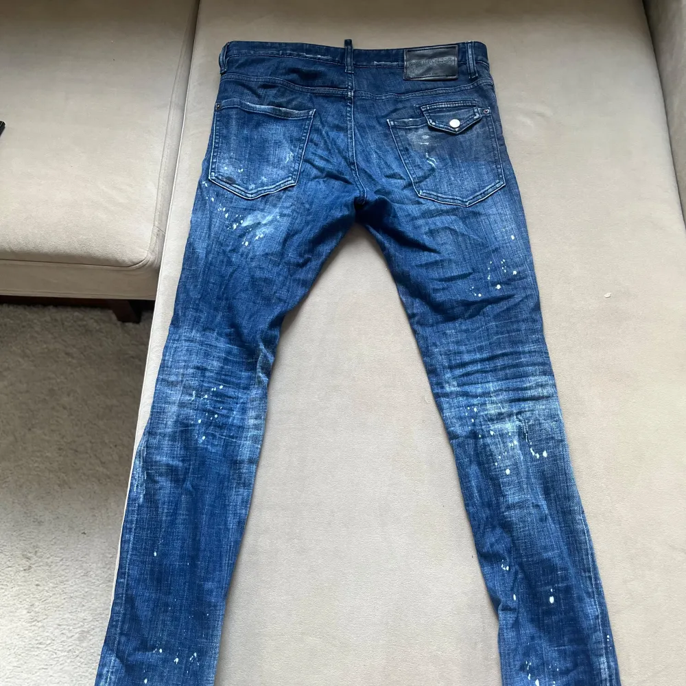 Säljer ett par snygga blå jeans från Dsquared2. De har en cool distressed look med slitningar och färgstänk. Perfekta för en avslappnad och trendig stil. Jeansen är i bra skick och har en normal passform. De är tillverkade i Italien och är av hög kvalitet. Nypris ca 6000. Jeans & Byxor.