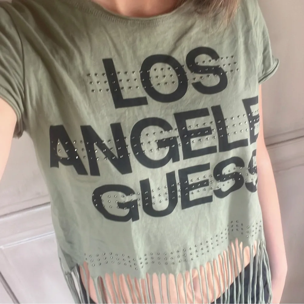 Säljer en cool olivgrön t-shirt från Guess med texten 'Los Angeles Guess' i svart. T-shirten är croppad med långa fransar längst ner. Lite stor i storleken.. T-shirts.