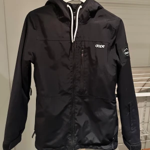 Svart vindjacka från Dope - Säljer en svart vindjacka från Dope med dragkedja och huva som är anpassad för skidåkare. Jackan har en stilren design med logotypdetaljer på bröstet och ärmen. Perfekt för blåsiga dagar och har en mönstrad insida för extra stil. Köptes för 1900kr❄️