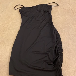 Svart axelbandslös klänning - Elegant svart axelbandslös klänning med smala axelband och rynkade detaljer på sidan. Perfekt för en stilren look. Från hm. Helt ny, har aldrig använt den. Passar för storlek s och xs!🫶🏼❤️