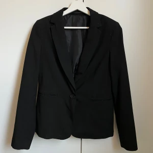 Svart kavaj - Elegant svart kavaj med klassisk design och enkelknäppning. Perfekt för en stilren look. Långärmad med smal passform och diskreta fickor framtill.