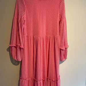 Rosa randig klänning från Vero Moda - Charmig rosa klänning från Vero Moda med vita ränder. Klänningen har trekvartsärmar och volangdetaljer vid ärmslut och nederkant. Perfekt för en somrig look!