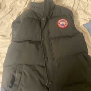 Svart dunväst med logotyp - Säljer en svart dunväst från Canada Goose med en hög krage och knappar framtill. Västen har en broderad logotyp på bröstet och är perfekt för kyligare väder.