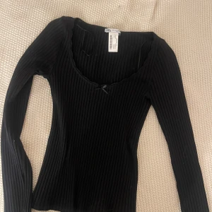Svart ribbad topp från Zara - Snygg svart ribbad topp från Zara med långärmad design och en liten rosett framtill. Perfekt för en stilren look.