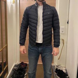 Svart pufferjacka från Gant - Säljer en stilren svart pufferjacka från Gant. Jackan har en dragkedja framtill och är långärmad. Perfekt för kyligare dagar med sin quiltade design som håller värmen.