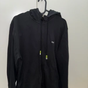 Svart huvtröja från Boss - Snygg svart huvtröja från Boss med dragkedja och justerbar huva. Tröjan har långa ärmar och en diskret logga på bröstet. Perfekt för en avslappnad stil.