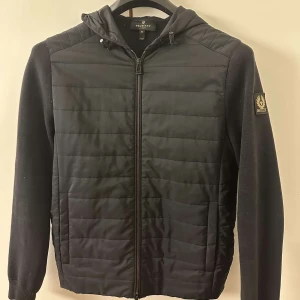 Svart jacka från Belstaff - Snygg svart jacka från Belstaff med quiltad framsida och huva. Jackan har dragkedja och ett stilrent märke på ärmen. Perfekt för en cool och avslappnad stil. Nypris 3500kr/ pris kan diskuteras.