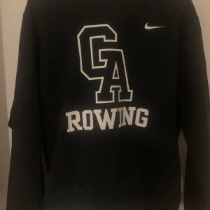 Svart tröja från Nike med tryck - Säljer en svart tröja från Nike med trycket 'GA Rowing' på framsidan. Tröjan har långa ärmar och är perfekt för en sportig look. Klassisk Nike-stil med en bekväm passform.