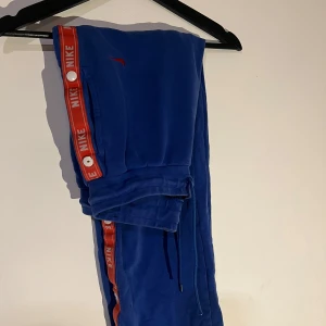 Blå mjukisbyxor från Nike - Säljer ett par blå mjukisbyxor från Nike med röd och vit logotyp längs sidorna. Byxorna har snörning i midjan och är perfekta för en avslappnad stil eller för ett träningspass. Byxorna har även knappar på sidorna som kan öppnas.