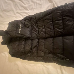 Svart dunväst från Sail Racing - Snygg svart dunväst från Sail Racing med dragkedja och hög krage. Perfekt för kyligare dagar och har en stilren design med logga på bröstet.