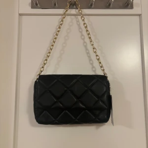 Svart quiltad axelväska med kedja - Snygg svart axelväska i quiltat mönster med en elegant guldkedja som axelrem. Perfekt för att ge en stilren touch till din outfit. Väskan har en praktisk storlek för att bära med sig det viktigaste.