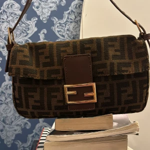 Mönstrad axelväska från Fendi - Snygg axelväska från Fendi med ikoniskt mönster i brunt och svart. Väskan har en justerbar axelrem och ett elegant spänne framtill. Perfekt för att ge din outfit en lyxig touch.