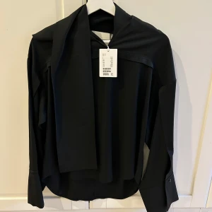 Svart ny blus med knytband från &otherstories - Elegant oanvänd svart blus med långa ärmar och knytband vid halsen från &otherstories.  Perfekt för en stilren look. Blusen har en lös passform och knappar vid ärmsluten. Köptes för 1200 kr 