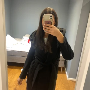 Svart kappa med huva - Snygg svart kappa med huva och knytband i midjan. Perfekt för kyliga dagar och ger en stilren look. Långärmad design som passar bra till både vardag och speciella tillfällen.