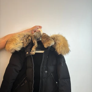 Svart dunjacka med pälsdetaljer från Cedrico - Snygg svart dunjacka från Cedrico med en fluffig pälsdetalj på huvan. Jackan har dragkedja och knappar framtill för extra skydd mot kylan. Perfekt för kalla vinterdagar.