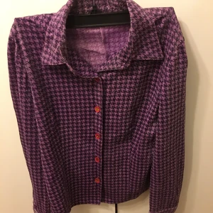 Lila och svart mönstrad skjorta - Snygg lila och svart skjorta med ett distinkt mönster och röda knappar. Perfekt för att sticka ut med stil. Skjortan har en klassisk krage och långa ärmar.