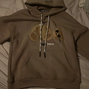 Beige hoodie från Palm Angels - Säljer en snygg beige hoodie från Palm Angels med broderade detaljer på framsidan. Hoodien har en klassisk känguruficka och justerbar huva med snören. Perfekt för en avslappnad stil.