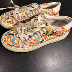 Tory Burch sneakers med geometriskt mönster - Säljer ett par unika Tory Burch sneakers med ett färgglatt geometriskt mönster i rött, blått, gult och orange. Skorna har snörning och en bekväm sula, perfekta för att liva upp vilken outfit som helst.