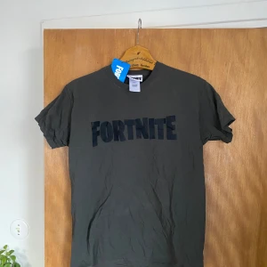 Grå Fortnite t-shirt - Säljer en grå t-shirt med Fortnite-logga i svart. Tillverkad i 100% bomull för en skön och bekväm känsla. Perfekt för alla Fortnite-fans! 👾Passar som S