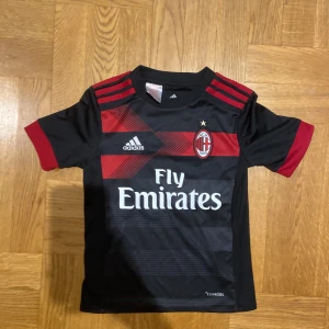 Snygg AC Milan tröja - Snygg AC Milan fotbollströja i svart och rött från Adidas. Den är designad med röda ränder på axlarna och Fly Emirates som sponsorlogga. Perfekt för alla Milan-fans!