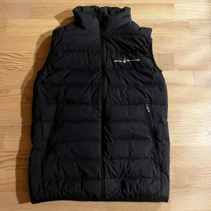 Svart dunväst från Sail Racing - Snygg svart dunväst från Sail Racing med dragkedja och hög krage. Västen är vindtät och andas bra, perfekt för kyliga dagar. Den har två praktiska fickor med dragkedja framtill. Storlek S/XS så passar super för lite mindre personer.