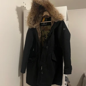 Svart parkajacka med pälsdetaljer från Blonde nr 8 - Snygg svart parkajacka från Blonde. Nr8  med pälsfodrad huva och camouflagemönstrat foder. Jackan har knappar och dragkedja framtill för extra skydd mot kyla. Perfekt för kalla dagar!