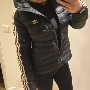 Svart dunjacka från Adidas - Snygg svart dunjacka från Adidas med de klassiska tre ränderna längs ärmarna. Jackan har en dragkedja framtill och en huva för extra skydd mot väder. Perfekt för kyligare dagar. Jackan har varit flitigt använd och säljer den därför billigt men den har dock inga skador. Den är storlek 36 men skulle rekommendera för 34. Kontakta för föer bilder och information🙌🏻
