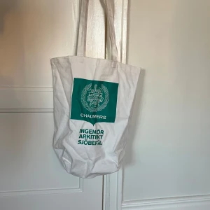 Vit tygväska med Chalmers-tryck - Säljer en vit tygväska med grönt Chalmers-tryck. Väskan har långa handtag och är perfekt för att bära böcker eller andra lätta föremål. Enkel och stilren design.