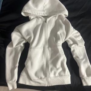Vit hoodie med dragkedja - Säljer en vit hoodie med dragkedja . Tröjan har långa ärmar och ribbade muddar vid ärmslut och nederkant.