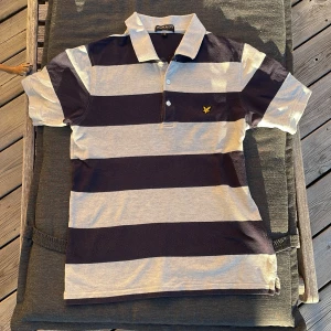 Randig pikétröja från Lyle & Scott - Säljer en snygg randig pikétröja från Lyle & Scott i storlek M men är liten som en XS. Tröjan har breda ränder i svart och grått med en klassisk krage och knappar framtill. Den är perfekt för både vardag och lite mer uppklädda tillfällen. Den har en broderad logga på bröstet som ger en extra touch av stil. Längd från axel 61 cm. Bred från bröstet 43 cm