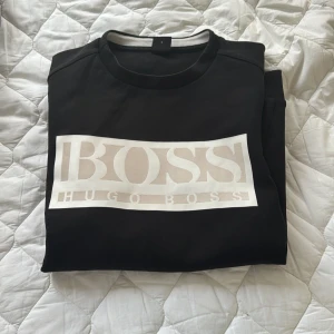 Svart tröja från Hugo Boss - Säljer en snygg svart tröja från Hugo Boss med deras ikoniska logga i vitt på framsidan. Tröjan har långa ärmar och är perfekt för en stilren look. Passar både till vardags och fest!