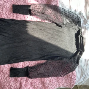 Svart klänning med transparenta ärmar - Säljer en elegant svart klänning med unika transparenta ärmar som har ett prickigt mönster. Klänningen är tight och har en stilren design som passar perfekt för festliga tillfällen. Den är långärmad och gjord i ett mjukt material som känns skönt mot huden.