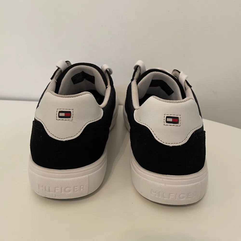 Säljer ett par snygga sneakers från Tommy Hilfiger i svart och vitt. Skorna har en klassisk design med vita snören och en vit sula. De är perfekta för vardagsbruk och har en stilren look med Tommy Hilfiger-loggan på sidan o. Skor.